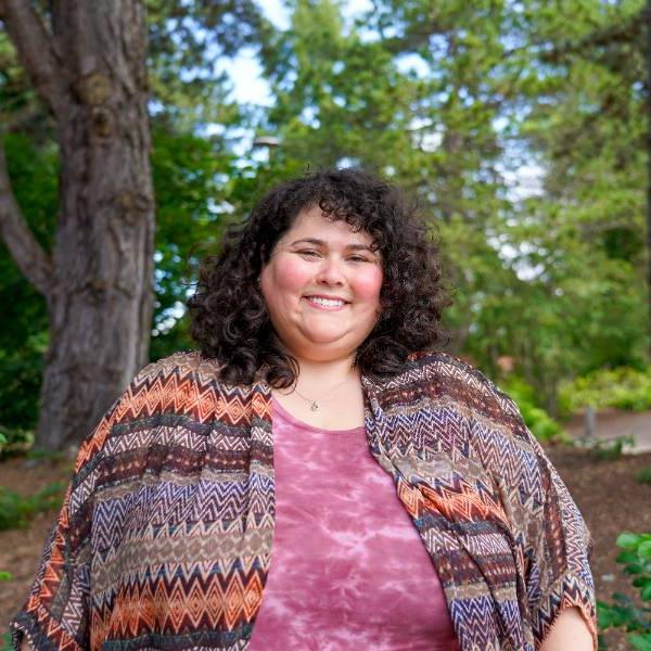 A headshot of Davi. She smiles against a wooded background, wearing a tie-dye pink shirt and patterned sweater.