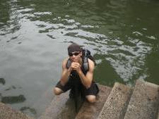 Amon Greene smiling, crouched down on steps leading to water with sunglasses and a backpack on. 